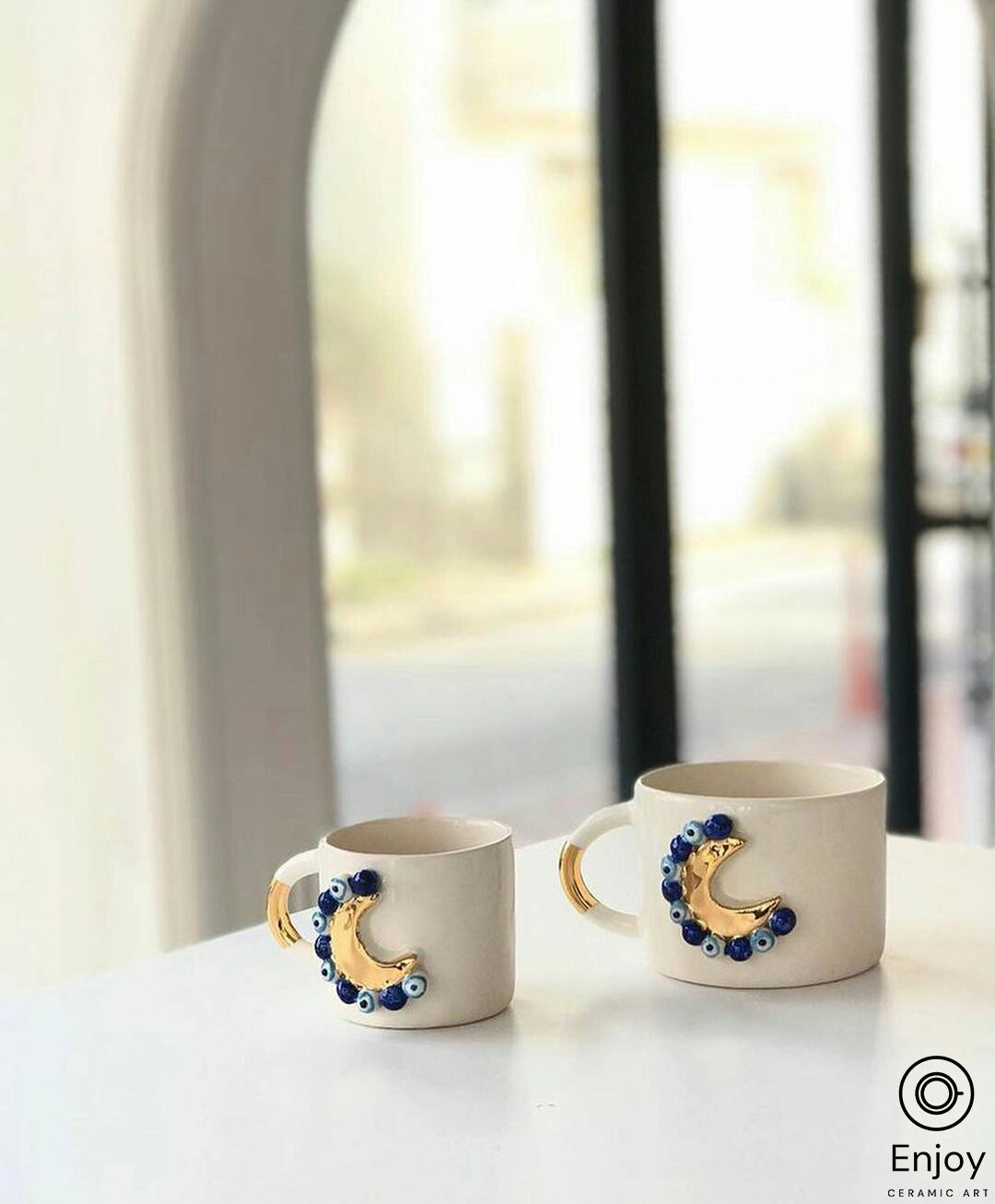 Two 'Blue Moon' ceramic mugs with gold handles and intricate blue beadwork on the crescent moon, elegantly set against the soft blur of a café window.