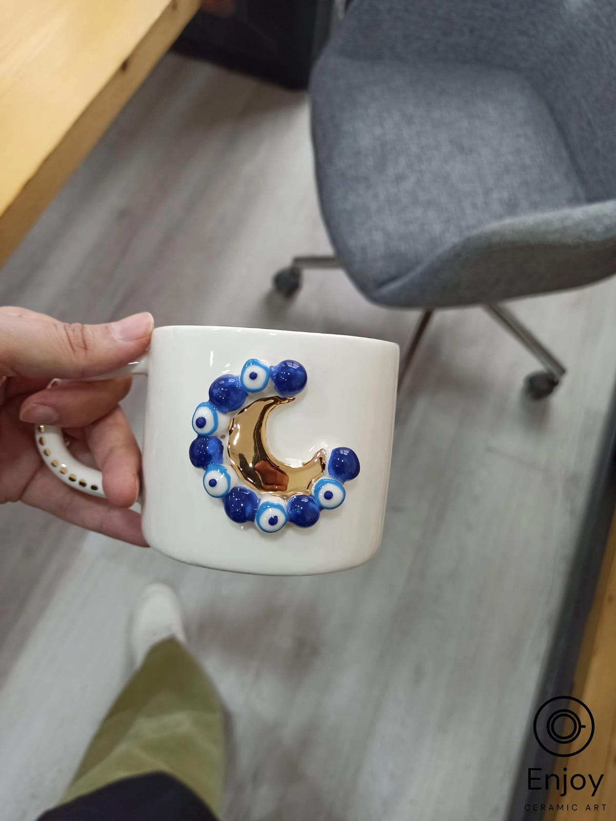 A hand holding a 'Blue Moon' ceramic mug, detailed with a golden crescent and blue evil eye beads, in an office setting with a gray chair and wooden desk in the background.