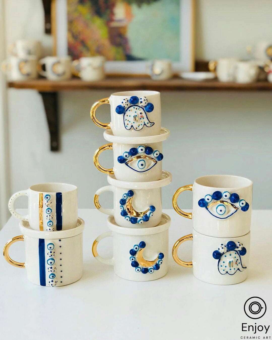 Stacked 'Blue Moon' ceramic mugs with gold handles, adorned with blue evil eye beads and crescent moon designs, displayed in a well-lit art studio.