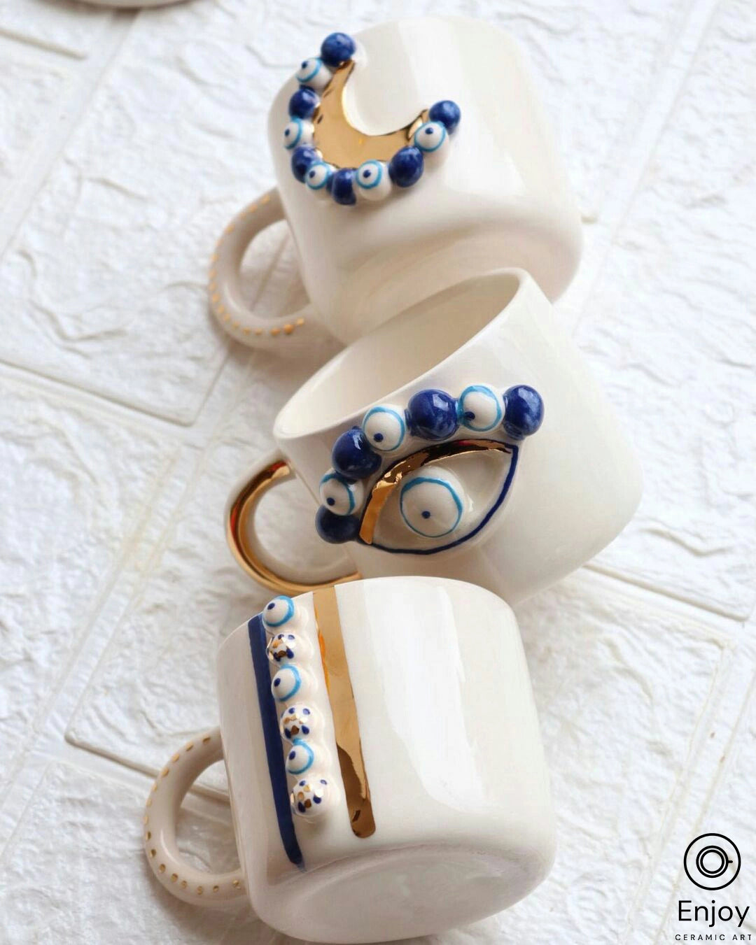 Three 'Blue Moon' ceramic mugs with gold handles and blue evil eye designs, artfully arranged on a textured white surface, showcasing the mugs' decorative elements.