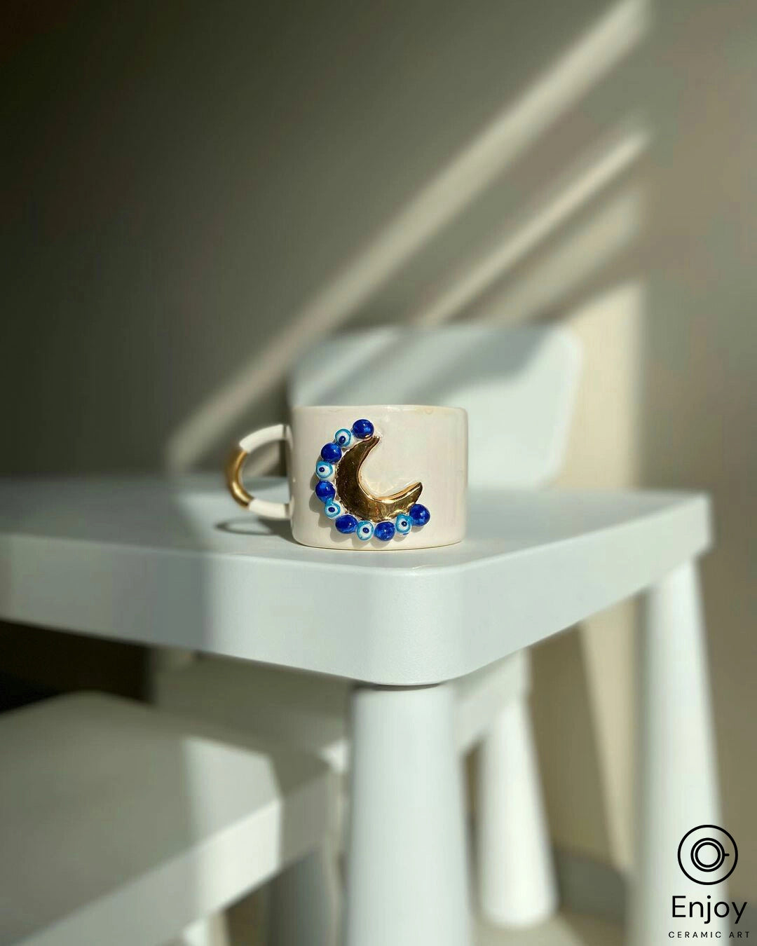 An artistic 'Blue Moon' ceramic mug with a golden crescent and blue evil eye beads on a modern white chair, bathed in soft sunlight casting geometric shadows.
