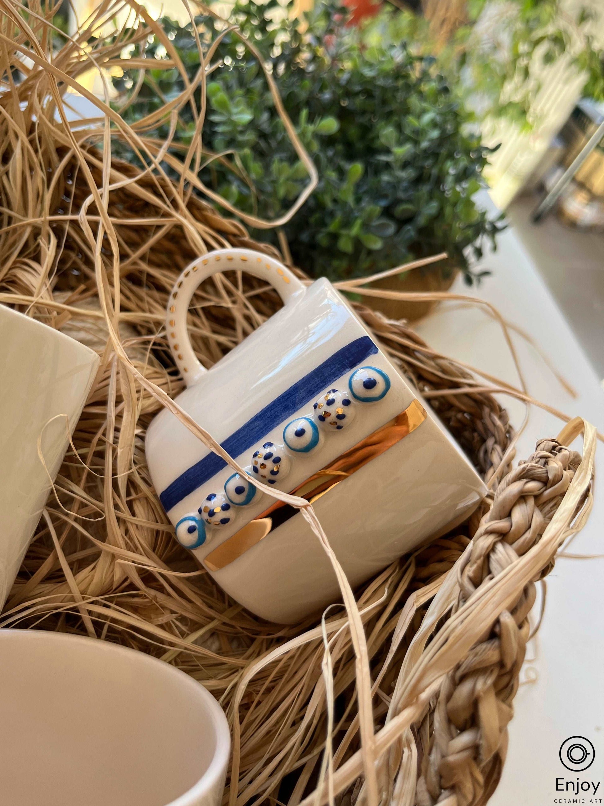 A white ceramic mug with a golden rim, adorned with blue evil eye patterns, partially hidden in a nest of natural straw, resting on a woven placemat, with a blurred background of green plants, presenting a cozy and inviting atmosphere.