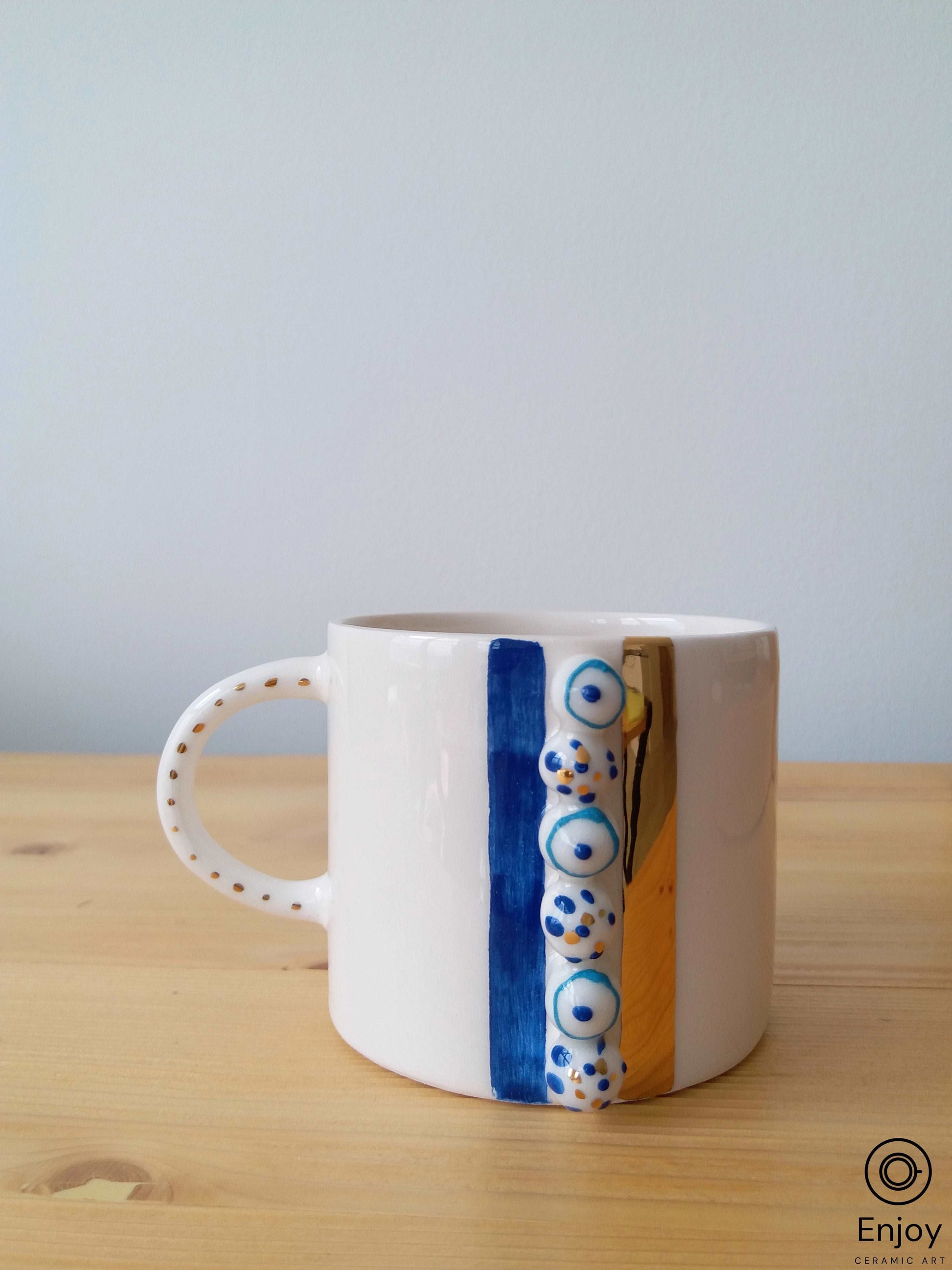A white ceramic mug with a polished golden handle, detailed with a vertical strip of blue and white evil eye motifs, standing on a wooden surface against a neutral light blue background.