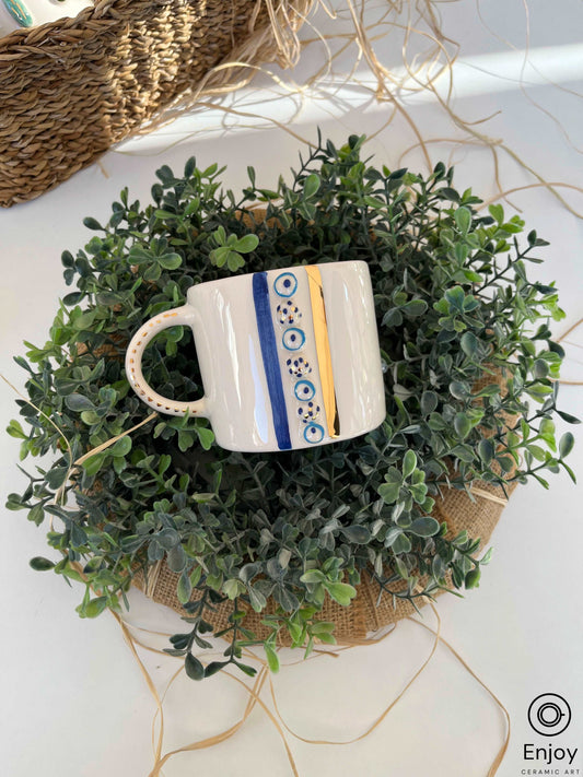 A white ceramic mug with a golden rim and handle, decorated with blue evil eye amulets, nestled in a lush green artificial plant wreath, set on a woven circular placemat with stray raffia strands, on a white surface.