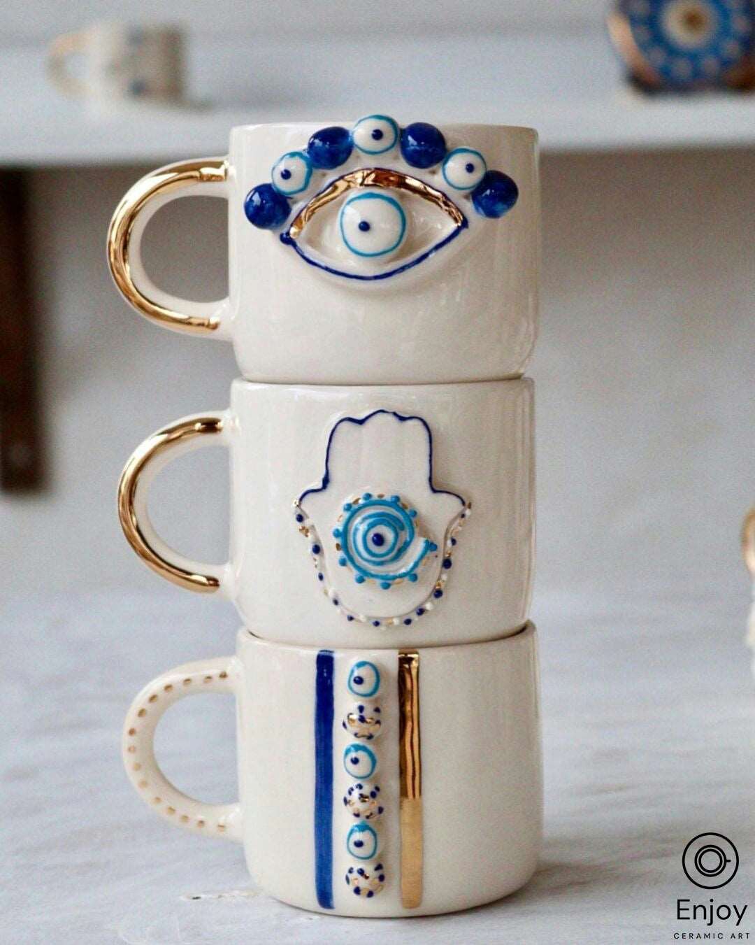 A stack of three white ceramic mugs with golden handles, each featuring a different blue evil eye design; the top with an eye, the middle with a hamsa hand, and the bottom with a stripe of multiple evil eyes, against a soft white background with light blue accents.