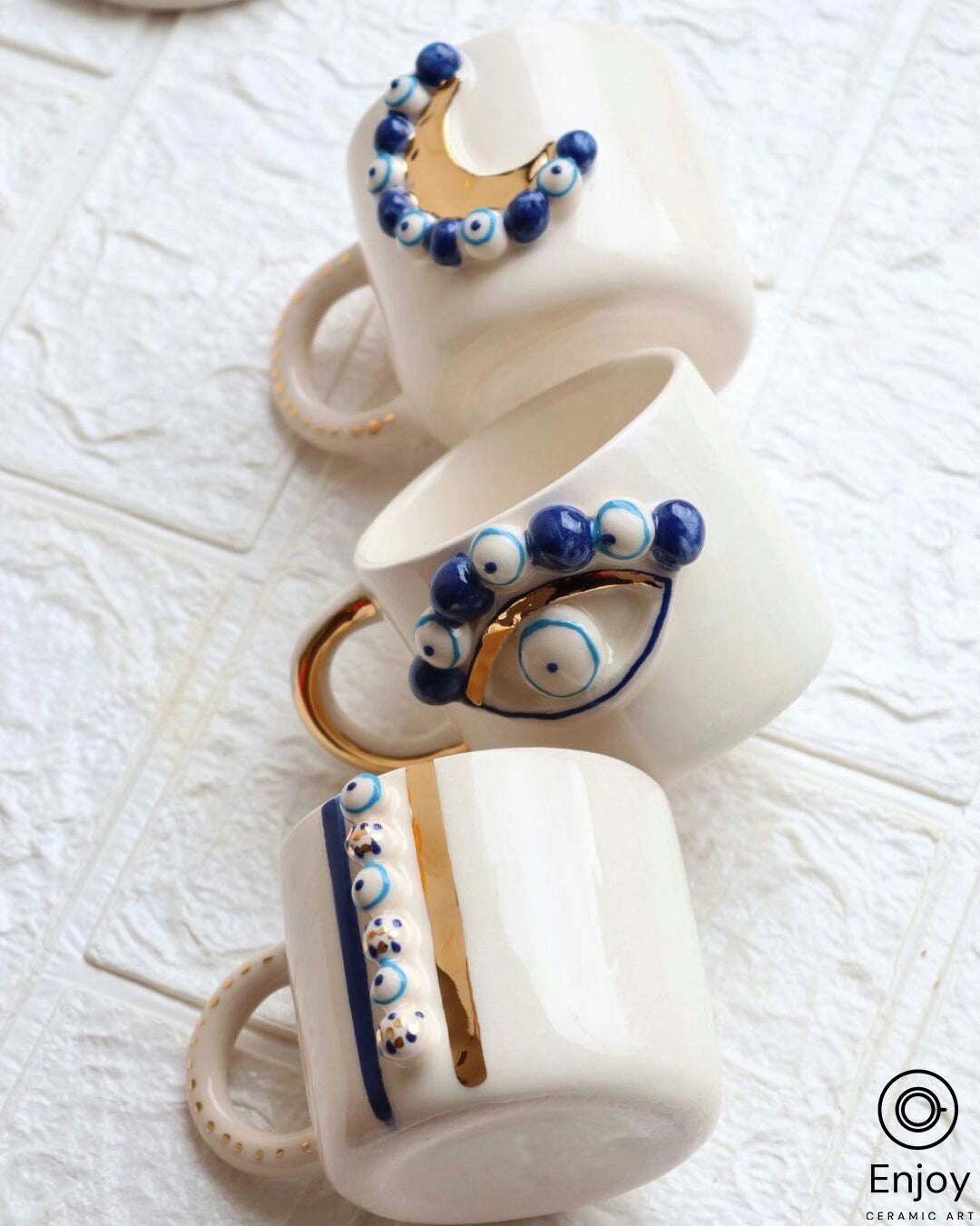 Three white ceramic mugs with golden details and blue evil eye designs, creatively stacked and angled to show each unique pattern, on a white textured surface evoking an elegant and artistic vibe.