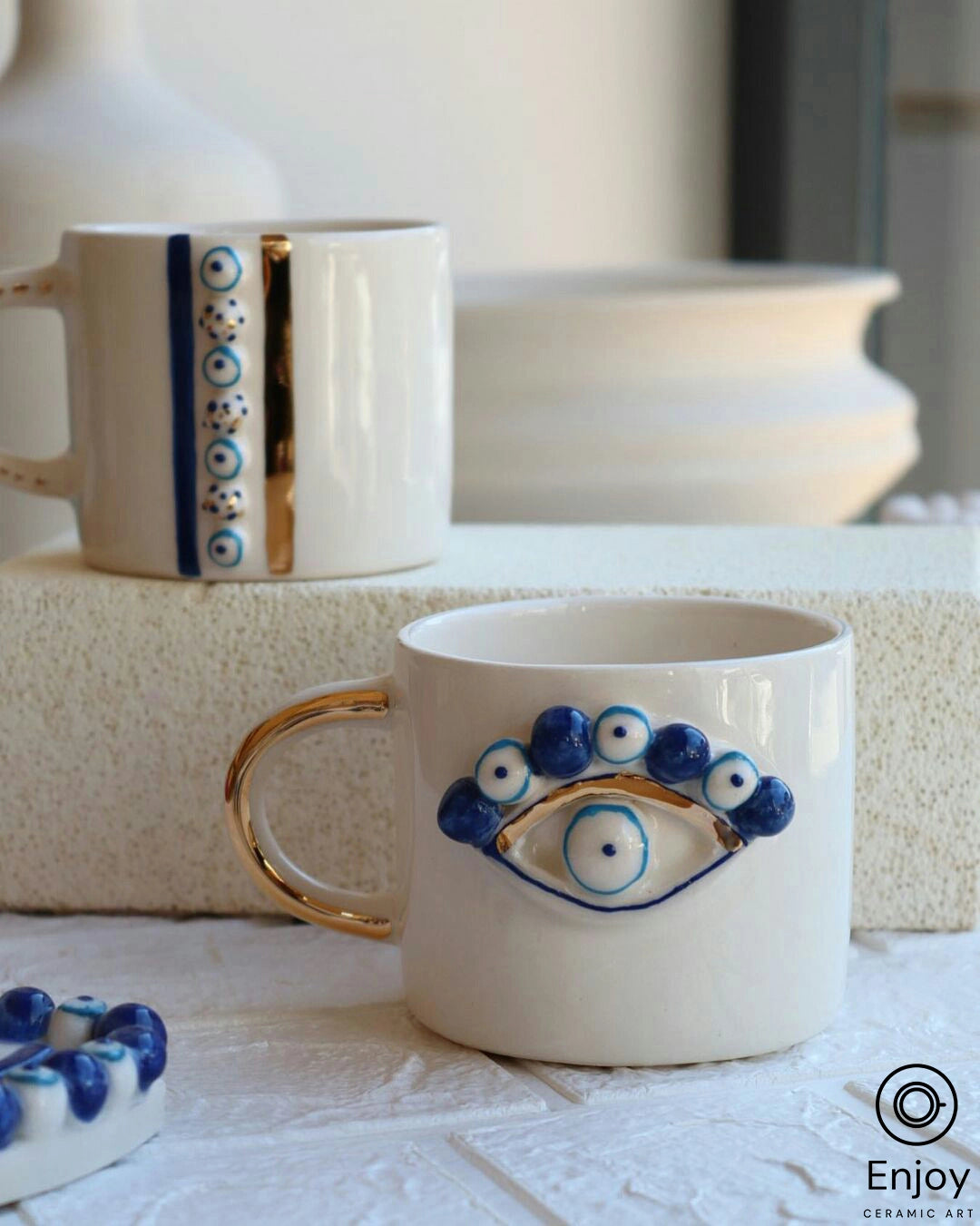 A prominent white ceramic mug with a glossy golden handle, featuring a large blue evil eye design on the side, situated on a marbled countertop with another similarly patterned item in the blurred background.