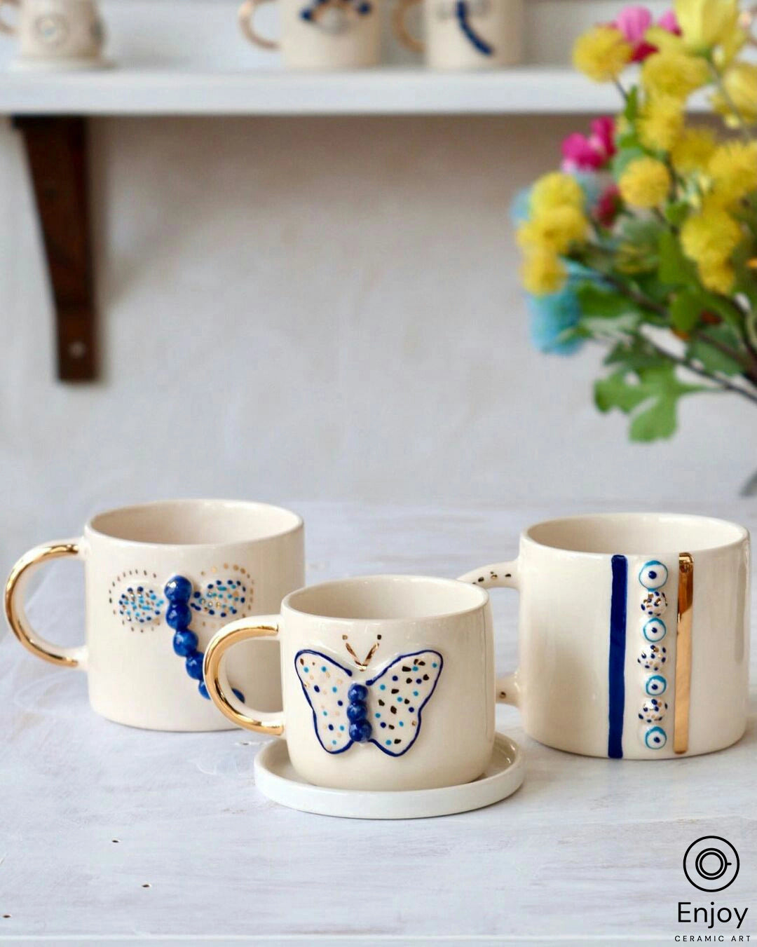 Three white ceramic mugs with golden handles on a white tabletop, each featuring unique blue designs: the left with evil eye beads, the center with a painted blue butterfly, and the right with a vertical line of evil eyes, displayed in front of a shelf with vibrant flowers adding a splash of color to the serene setting.