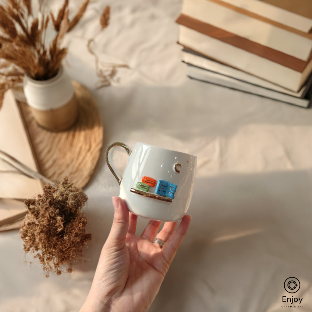 A hand holds a white mug with a 3D bookshelf design, surrounded by dried flowers and stacked books