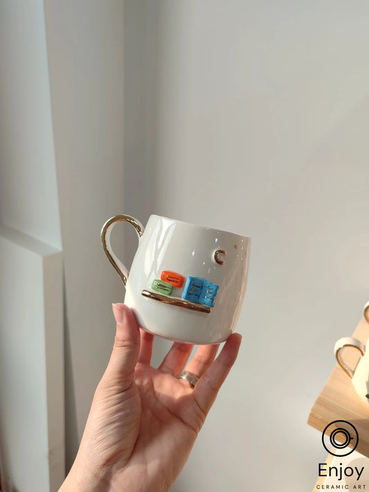 A hand holds a white mug with a golden handle, adorned with colorful book illustrations, against a soft-lit background.