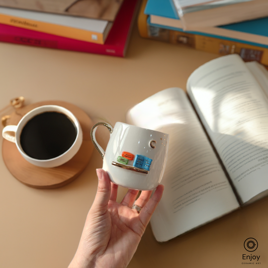 A white mug with a 3D bookshelf design is held above an open book and a cup of black coffee.