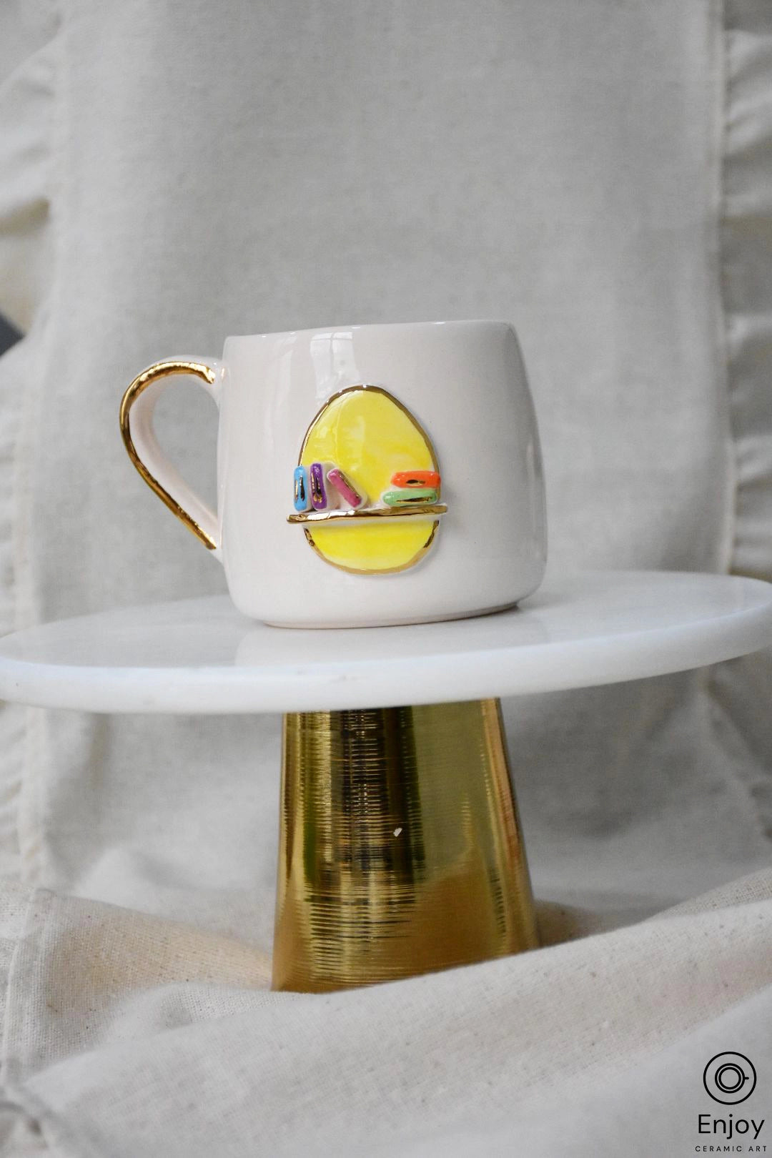 A chic white mug adorned with a colorful 3D bookshelf design, resting on a golden pedestal against a linen canvas.
