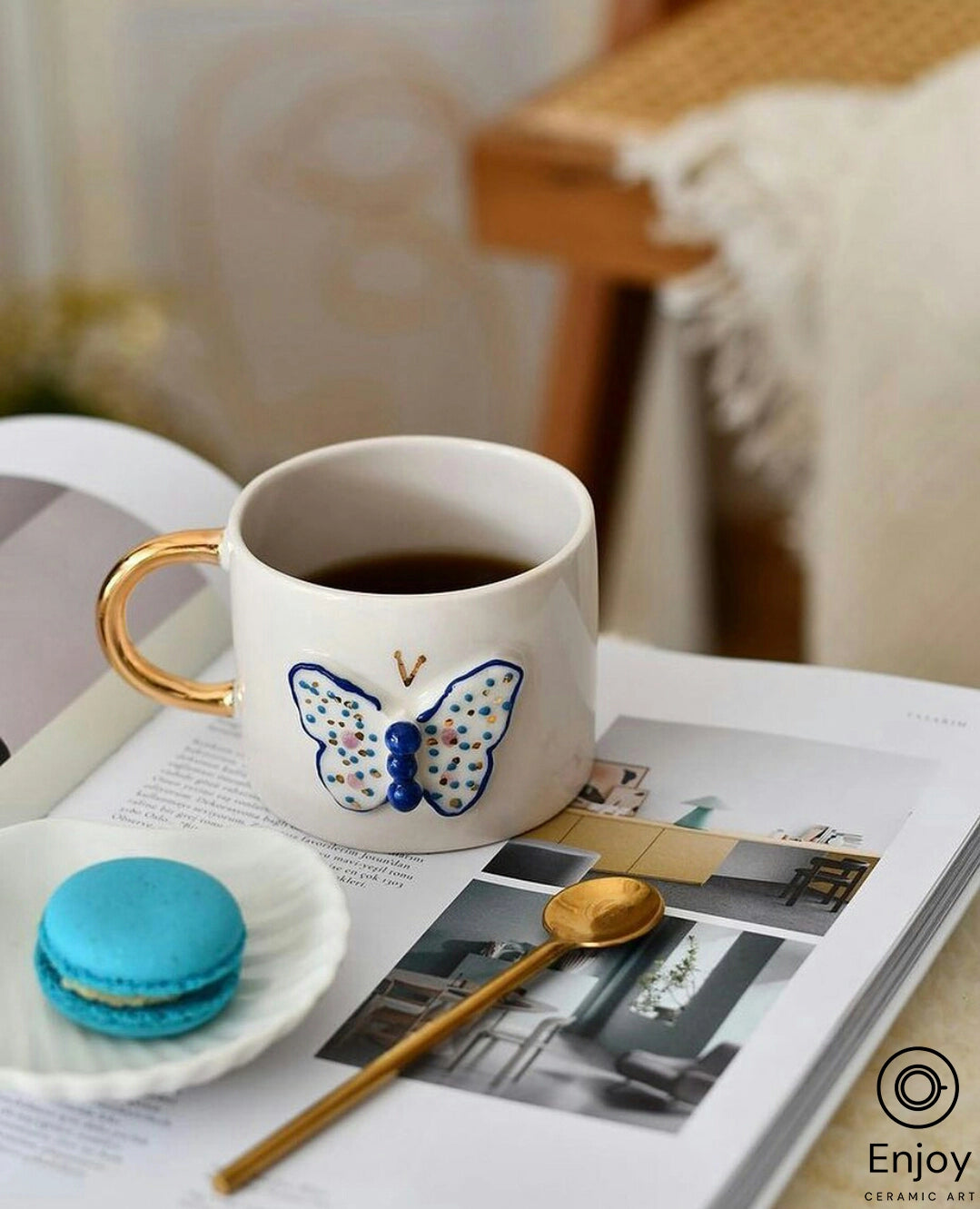 A white ceramic mug with a golden handle and a delicately painted butterfly motif, filled with dark liquid, stands on an open magazine featuring home decor. A single blue macaron and a golden spoon rest beside the mug, set against a cozy room backdrop with warm lighting