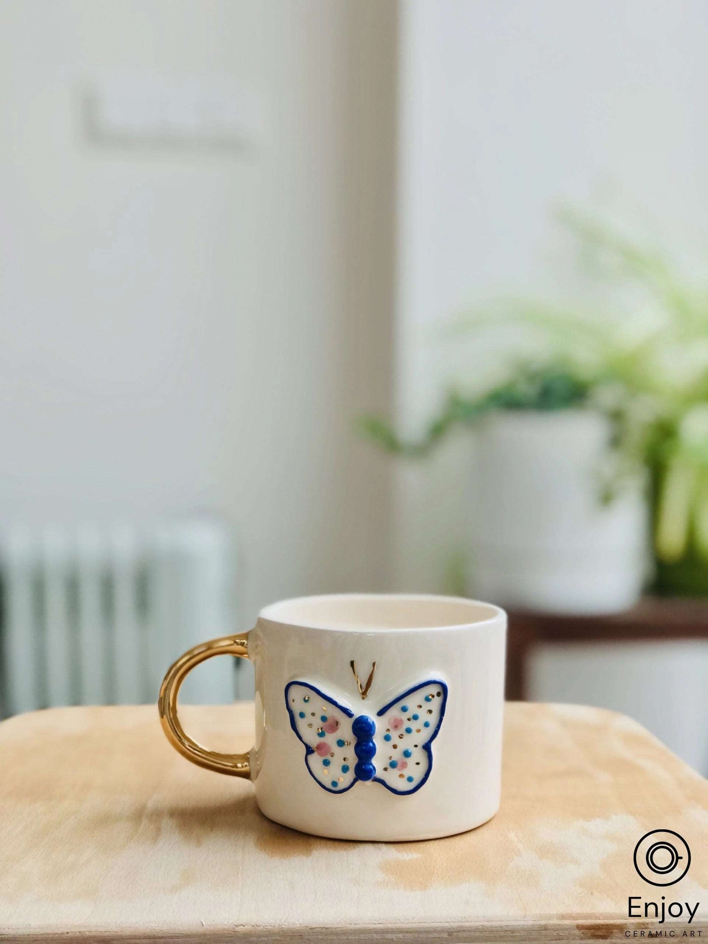 A white ceramic mug with a shiny golden handle and a hand-painted blue butterfly design, standing on a wooden table. In the soft-focus background, a green potted plant adds a touch of freshness to the tranquil setting.