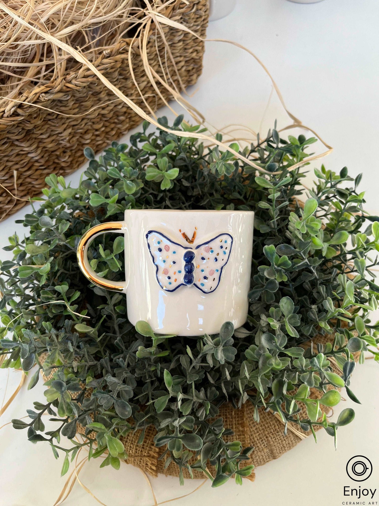 A white ceramic mug with a golden handle and a blue butterfly illustration is nestled in a lush bed of greenery, with a woven basket and dried straw adding texture to the composition, creating a harmonious blend of nature and artisanal craftsmanship