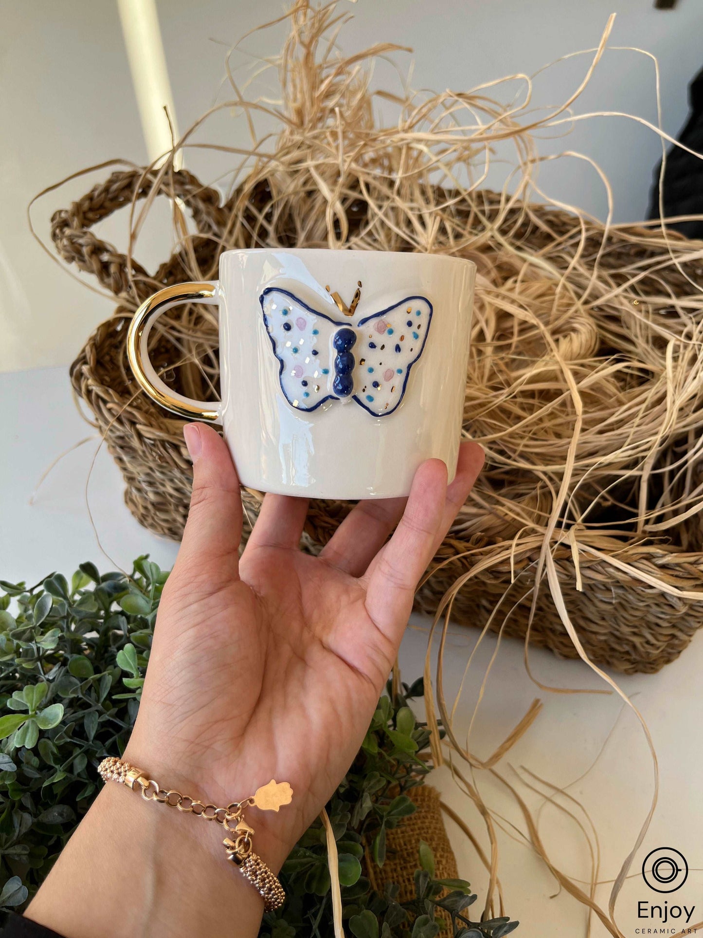 A hand holding a white ceramic mug with a gold handle and a painted blue butterfly, contrasted against a natural backdrop of dried straw and a rustic woven basket, evoking a cozy, artisanal vibe.