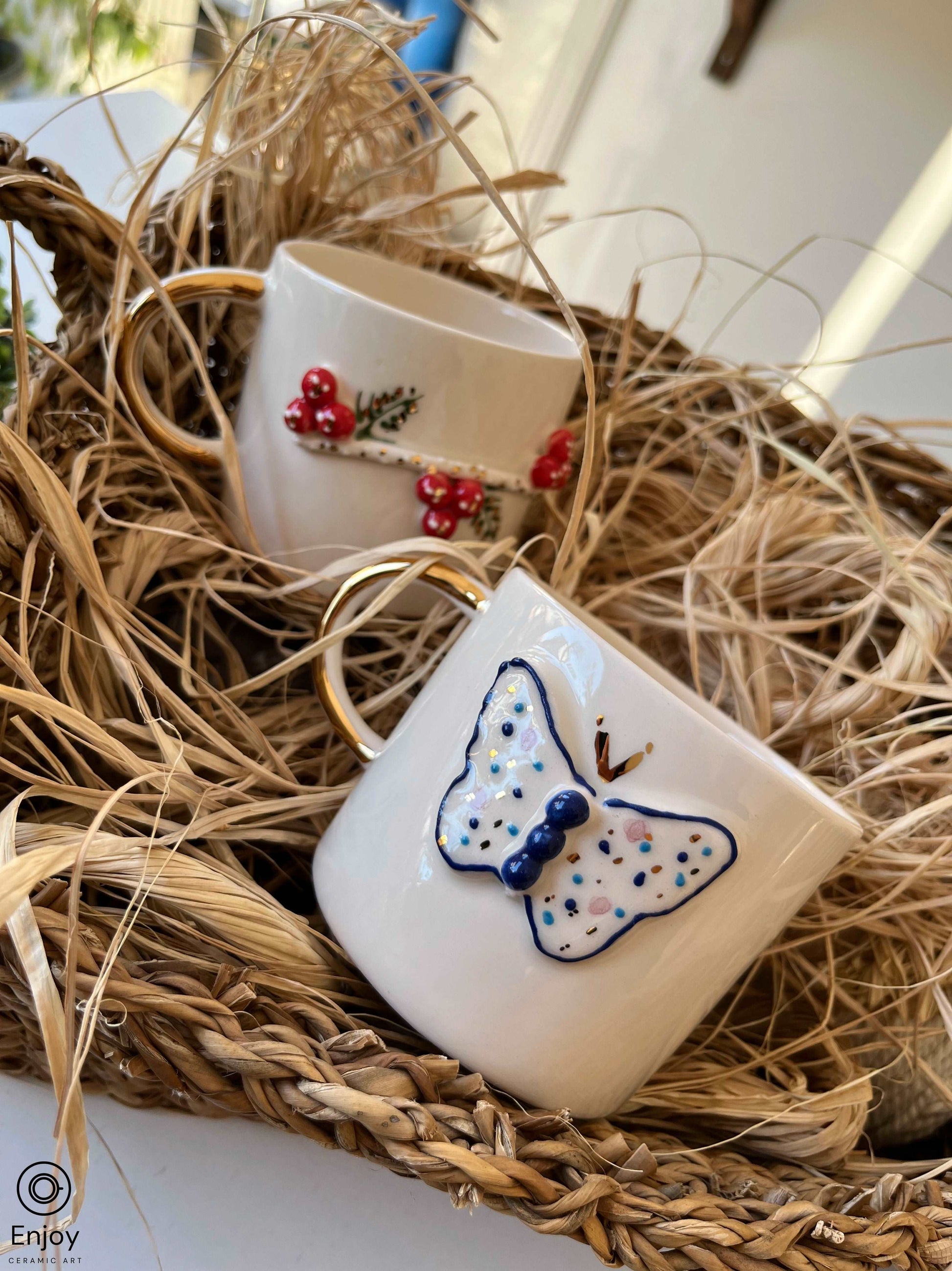 An intricately designed ceramic mug with a golden handle, nestled in a bed of straw within a woven basket. The mug features a delicate blue butterfly motif, adding a touch of whimsy to the rustic setting.