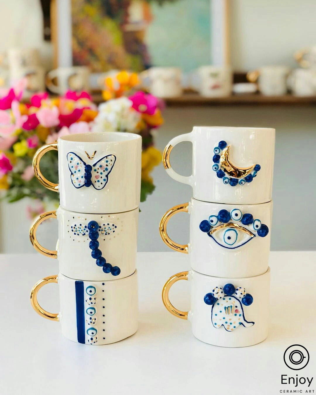A stack of four elegant white ceramic mugs with golden handles, each adorned with a different blue motif. The designs include a butterfly, an evil eye, and abstract dots, creating a charming and artistic display against a backdrop of vibrant flowers and a bright, airy room.