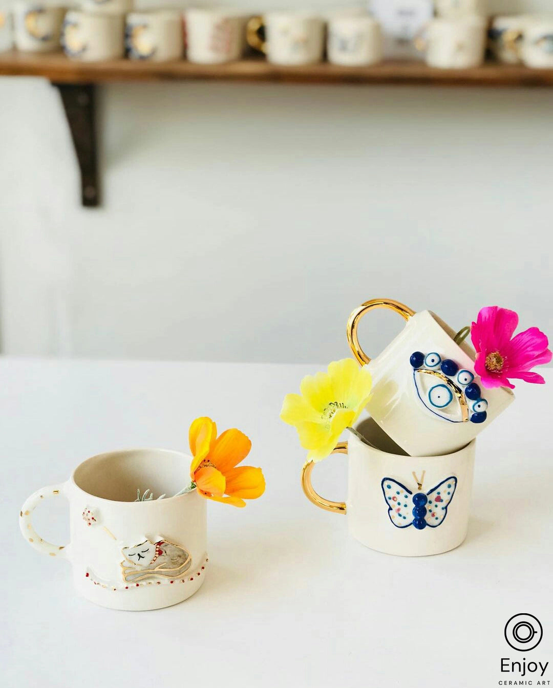 An artful arrangement of white ceramic mugs with golden handles, each featuring a unique blue design, such as an evil eye and a butterfly. Fresh flowers in vibrant orange, yellow, and pink hues are playfully placed in the mugs, adding a touch of natural beauty to the scene. In the soft-focus background, a wooden shelf holds an array of similarly styled mugs, contributing to the warm, creative atmosphere.