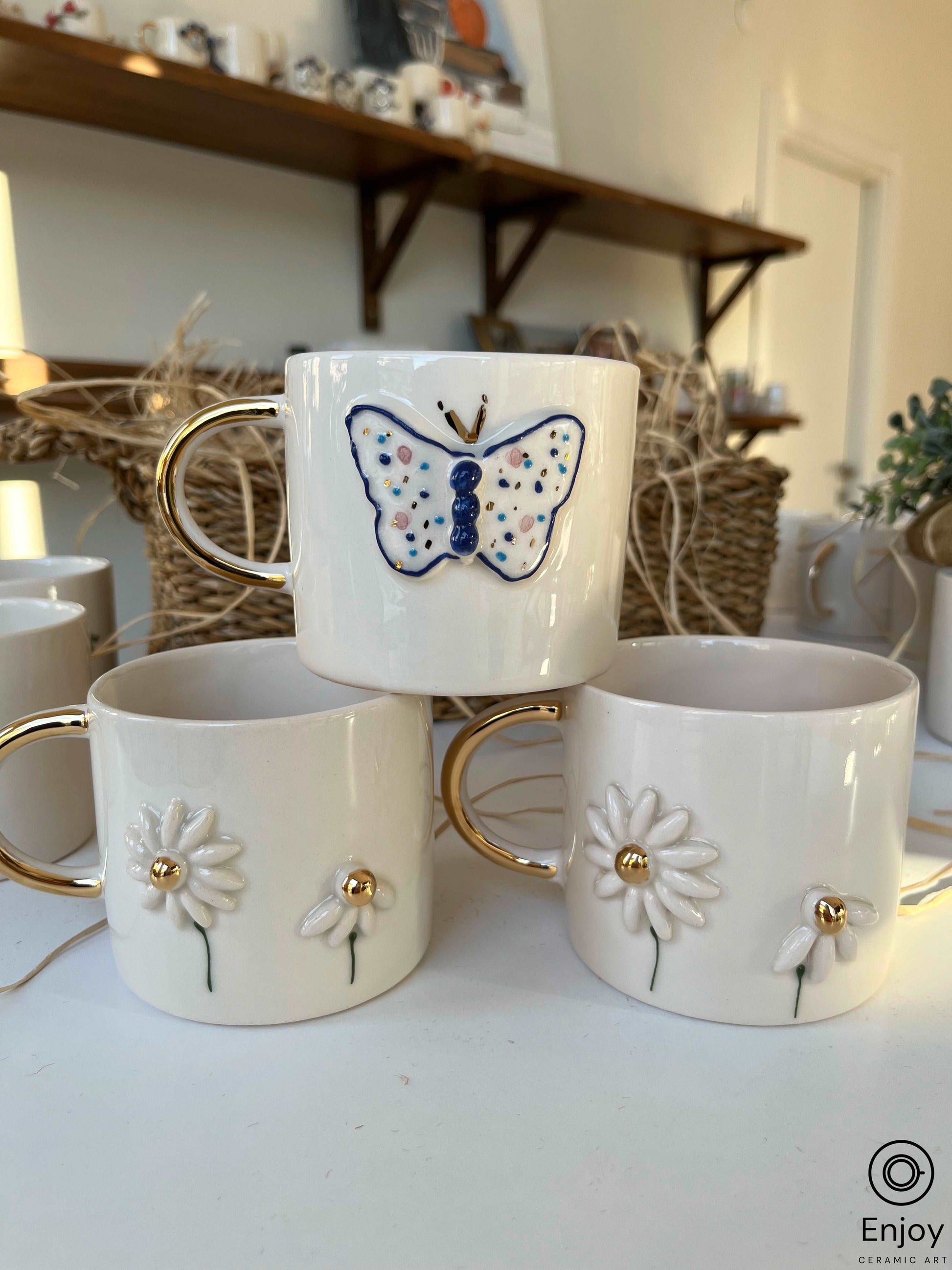 Three white ceramic mugs with golden handles, one with a blue butterfly, others with embossed daisies, against a straw basket backdrop.