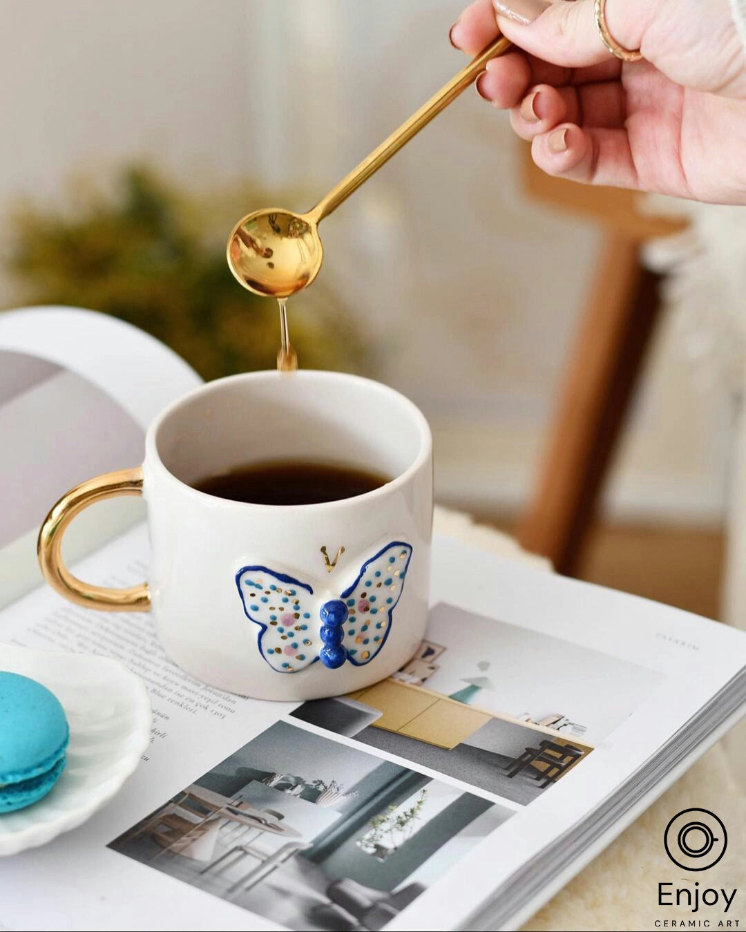 A hand is drizzling honey into a white ceramic mug with a golden handle and a blue butterfly design, which sits atop an open magazine with interior design images. The scene is accented by a blue macaron on the side, blending a moment of indulgence with a touch of elegance.