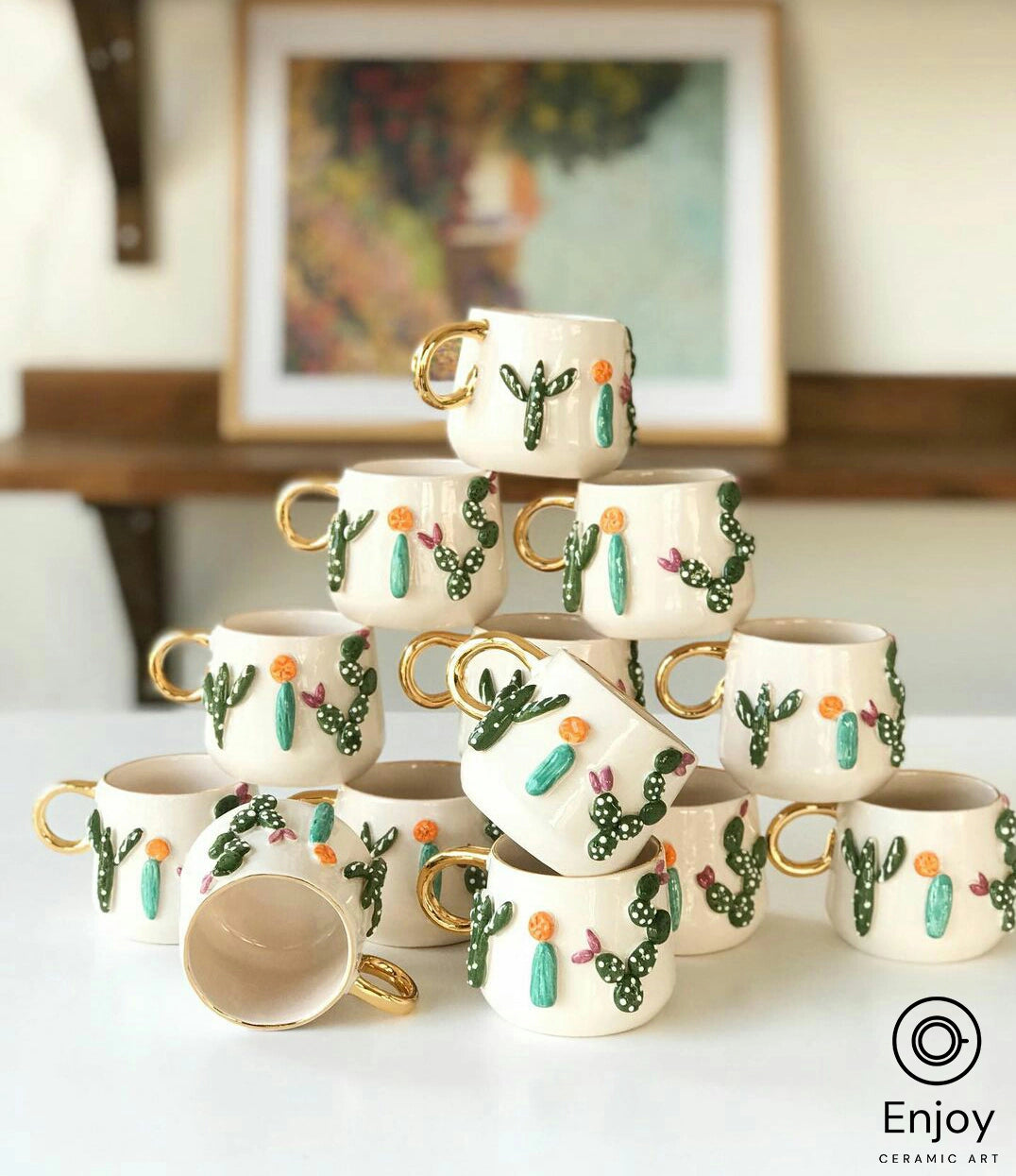 Stacked white mugs with golden handles and green cactus designs in an art studio, with a colorful painting in the background.
