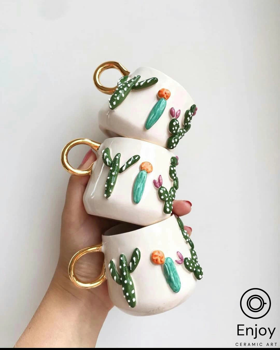 Hand holding three stacked white mugs with cactus designs and gold handles against a plain background.