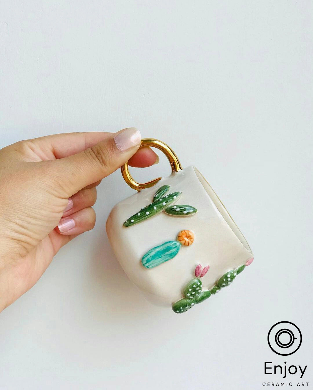 A hand holding a square-shaped white mug with a golden handle, adorned with green cactus designs and small colorful accents.