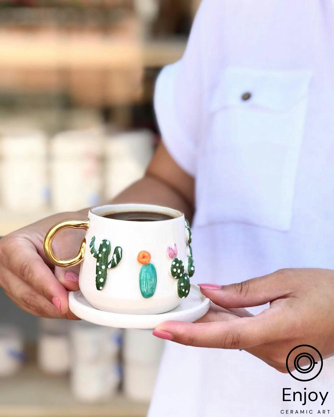 Two hands gently holding a white mug with a golden handle and a saucer. The mug is decorated with 3D cactus designs and colorful flowers, filled with coffee.
