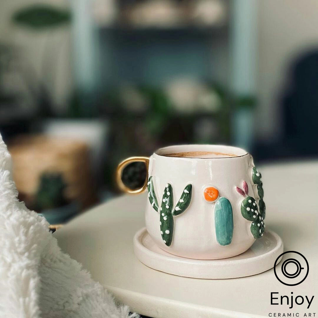A creamy white ceramic mug with a shiny golden handle, adorned with 3D cactus and flower designs, sits on a saucer filled with a frothy coffee beverage. The mug is set against a cozy indoor background.