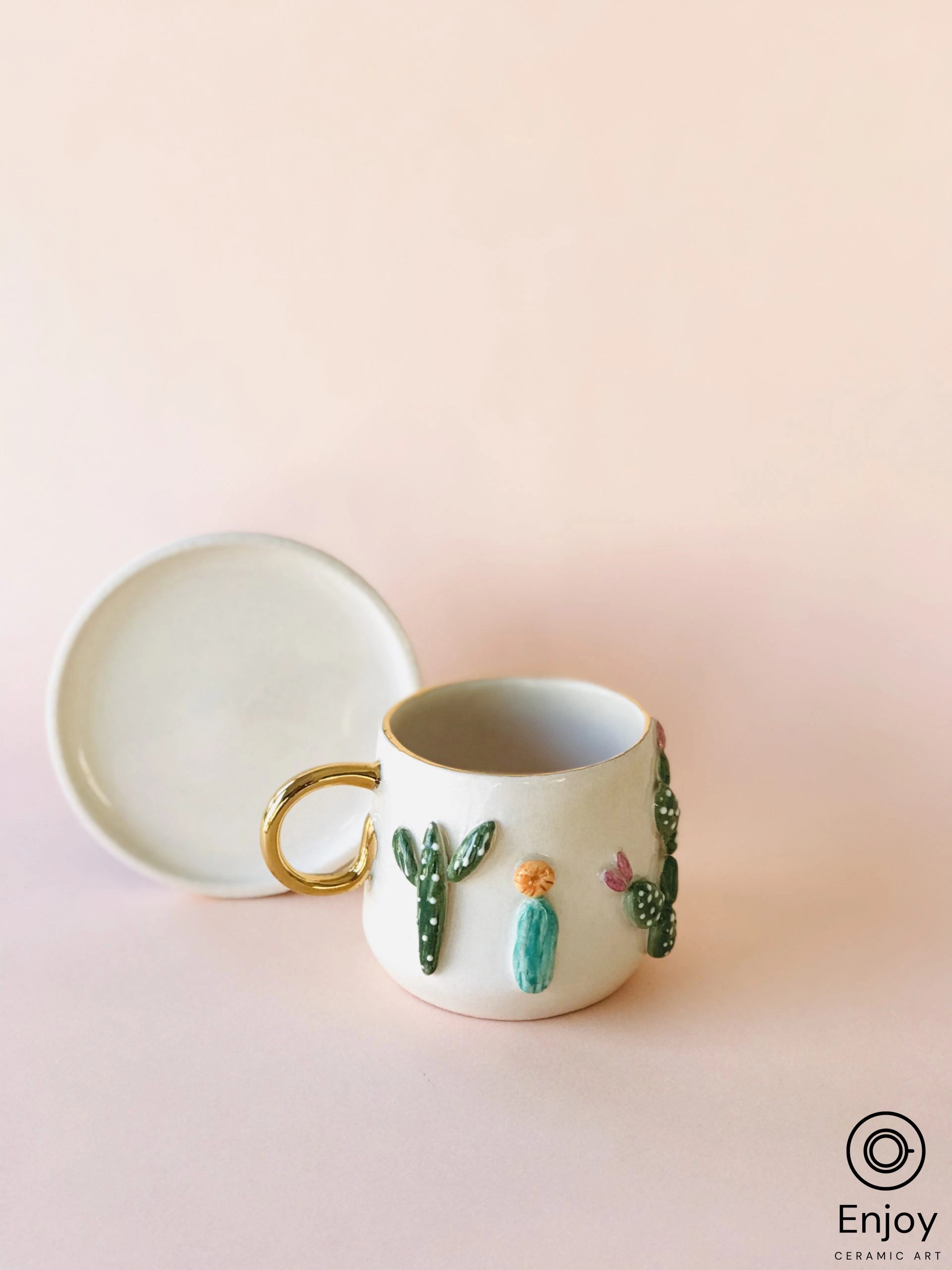 A ceramic mug with a gold handle, adorned with colorful 3D cactus motifs on a pastel pink background, conveys artistic and functional charm.