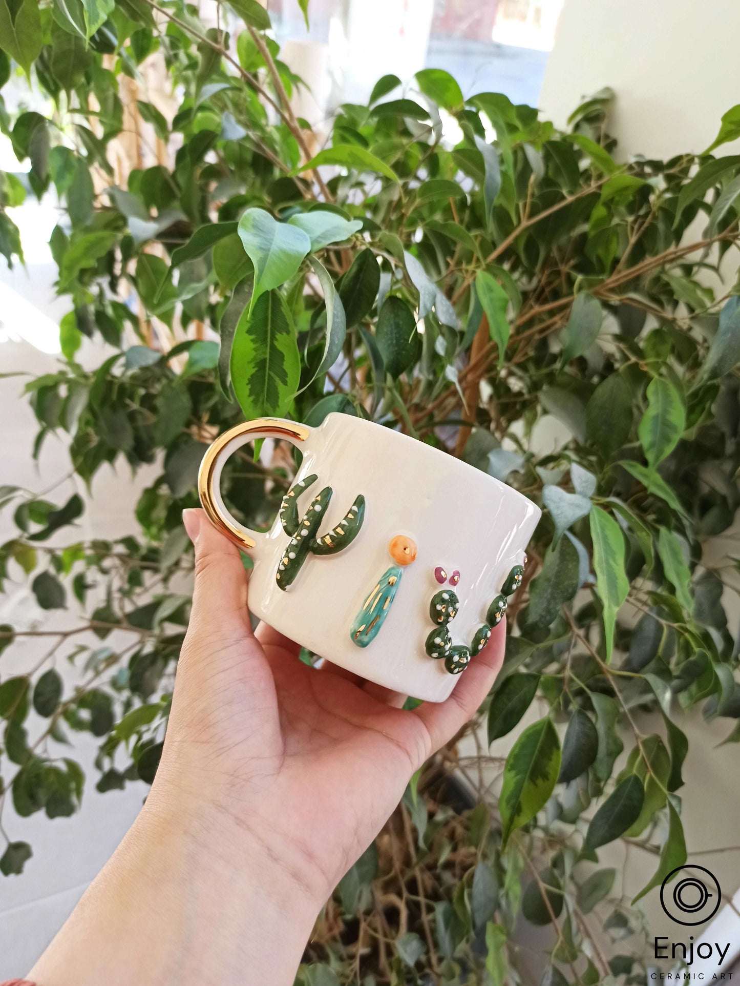 a hand holding a white ceramic mug adorned with three-dimensional cactus designs and a golden handle. It's set against a backdrop of leafy indoor plants, highlighting the mug's artistic nature-inspired embellishments.