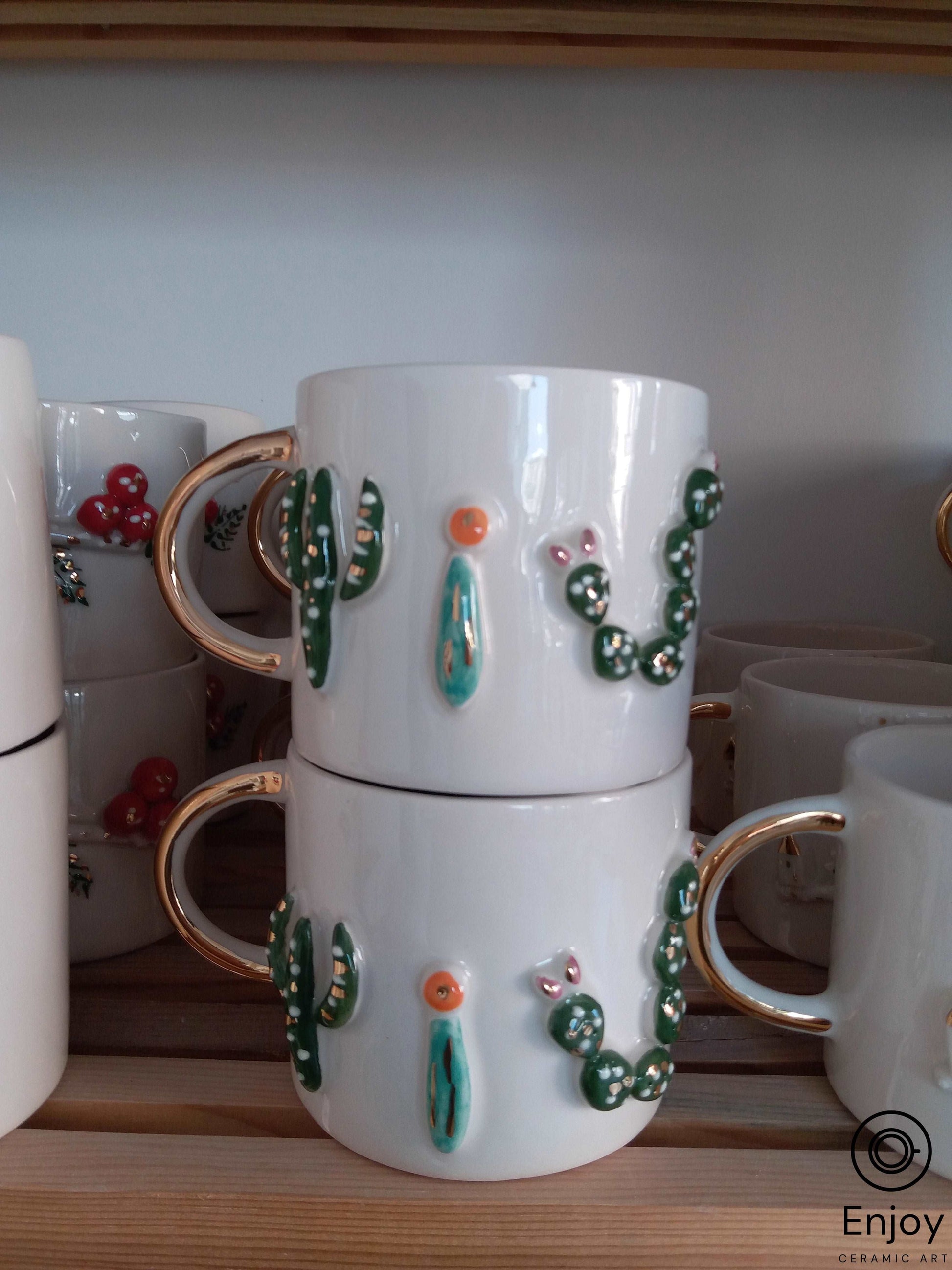 Stacked white mugs with gold handles, adorned with 3D cactus designs and tiny colorful blooms on a wooden shelf.