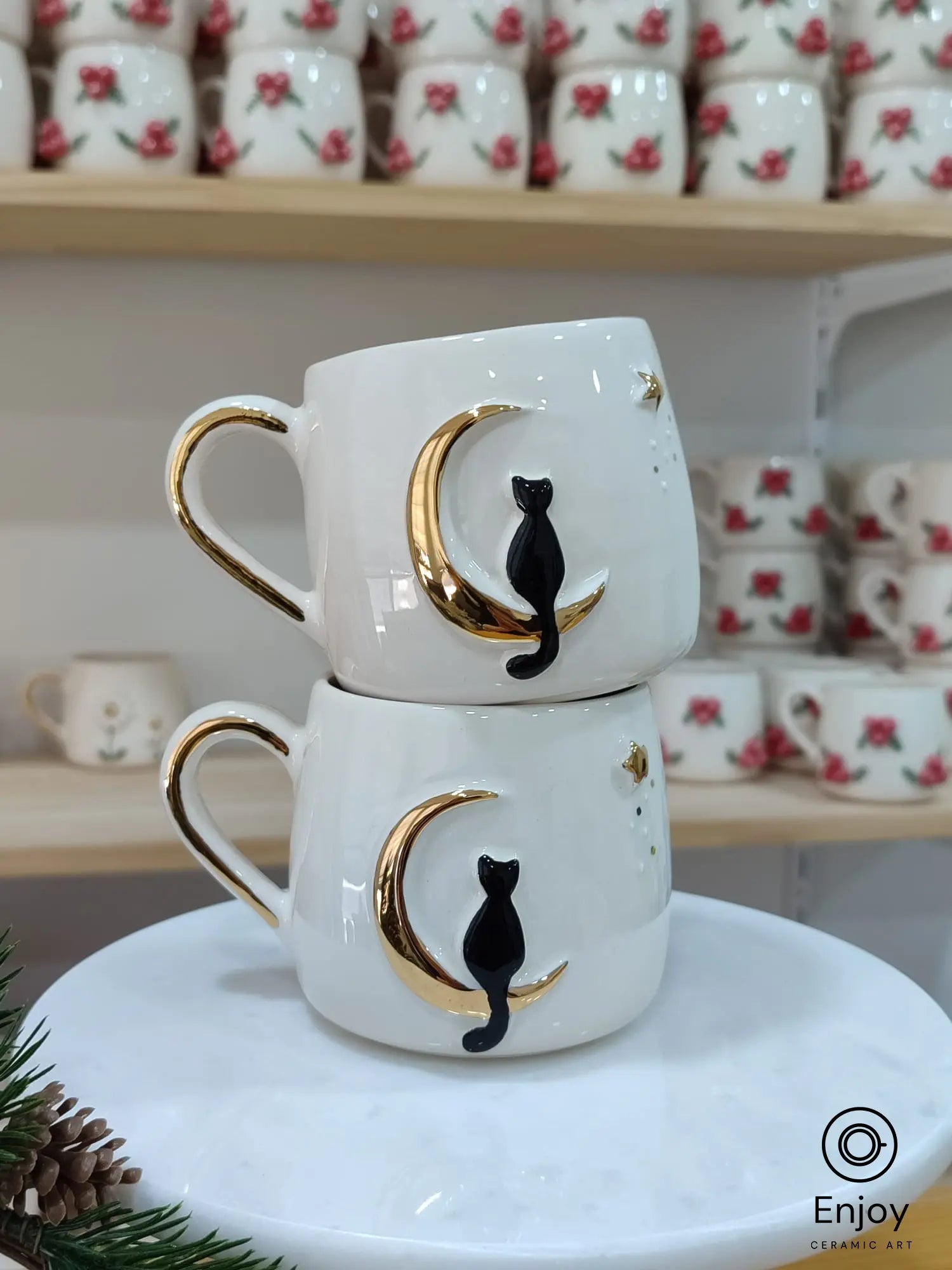 Handmade ceramic mugs featuring a black cat perched on a golden crescent moon, with a sleek gold handle. The mugs are stacked elegantly on a marble stand, with pinecones and festive décor surrounding them.