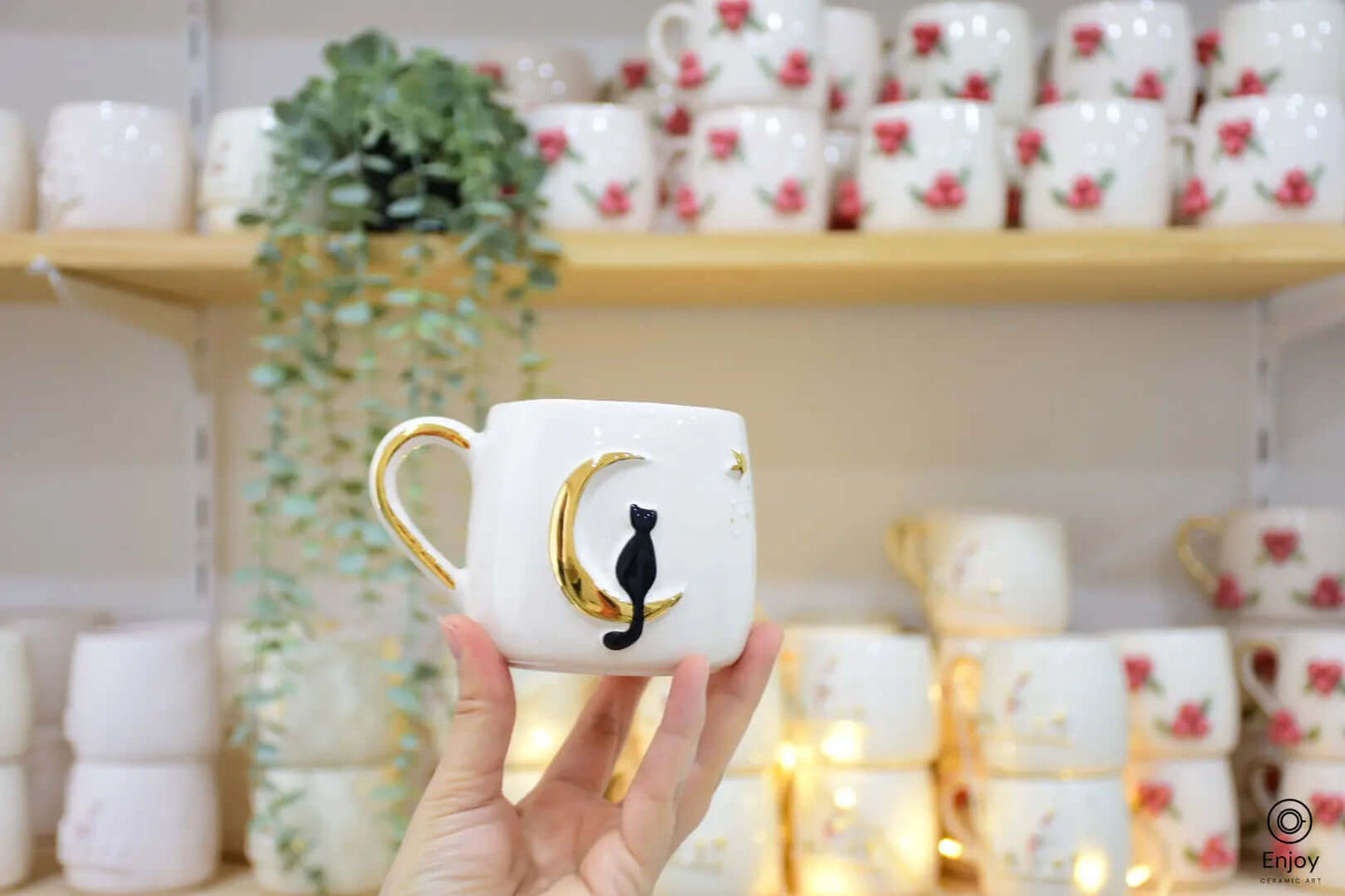 This image beautifully showcases the celestial cat mug being held against a cozy, warm background. The black cat sitting elegantly on a golden crescent moon exudes a charmingly whimsical vibe. The setting, adorned with fairy lights and surrounded by other handcrafted mugs featuring delicate floral designs, adds to the mug’s allure. This enchanting piece is perfect for anyone looking to add a magical touch to their coffee or tea rituals.