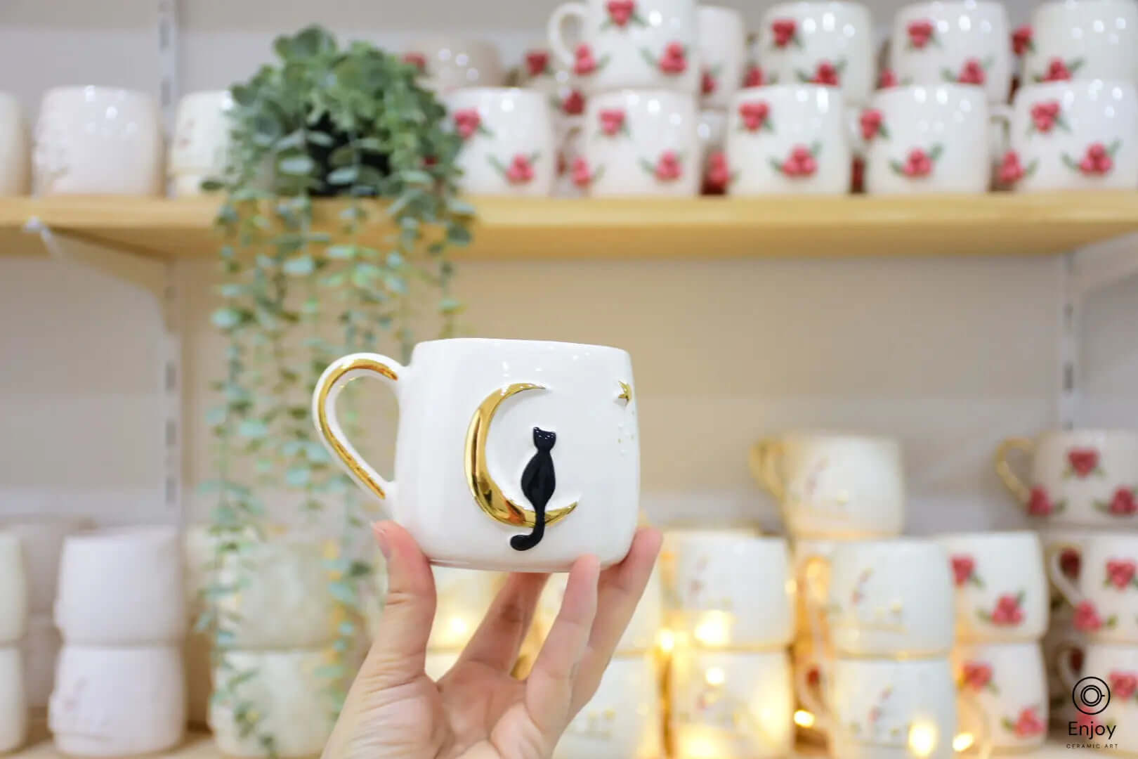 This image beautifully showcases the celestial cat mug being held against a cozy, warm background. The black cat sitting elegantly on a golden crescent moon exudes a charmingly whimsical vibe. The setting, adorned with fairy lights and surrounded by other handcrafted mugs featuring delicate floral designs, adds to the mug’s allure. This enchanting piece is perfect for anyone looking to add a magical touch to their coffee or tea rituals.