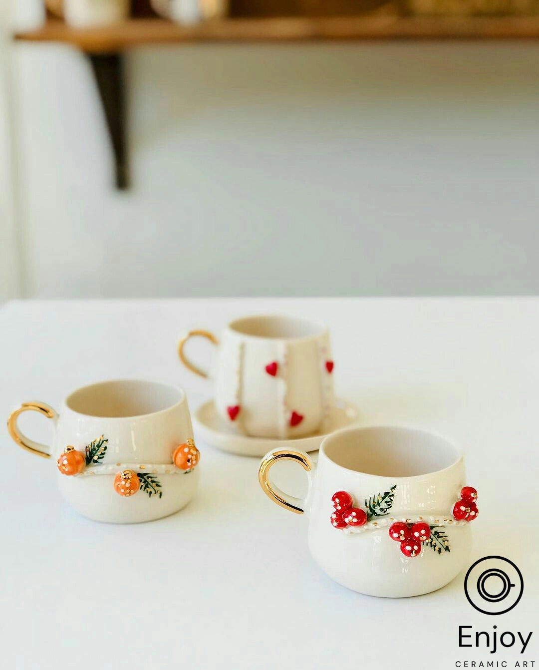 Three white ceramic mugs with gold handles, each adorned with a different festive design—pumpkins, hearts, and berries—on a white surface.