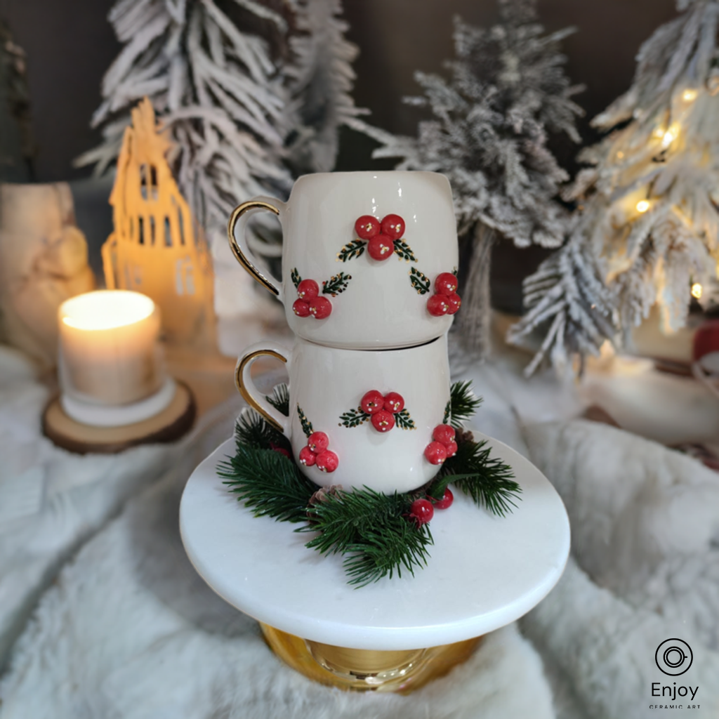 Two white mugs with holly berry designs, stacked on a gold pedestal, surrounded by festive winter decor with a candle and snow-covered trees in the background.