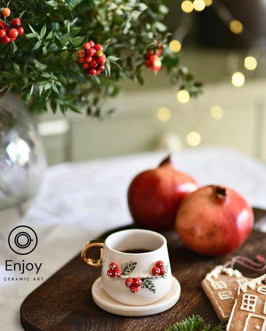 A festive white ceramic mug with a red berry and holly design, gold handle, accompanied by pomegranates and holiday decorations.
