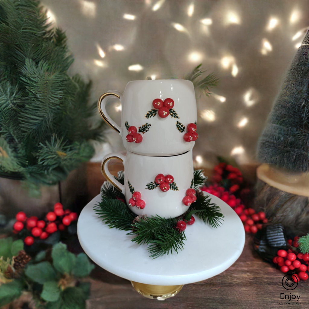 Two white mugs adorned with red holly berries stacked on a gold pedestal, surrounded by festive greenery and holiday lights.