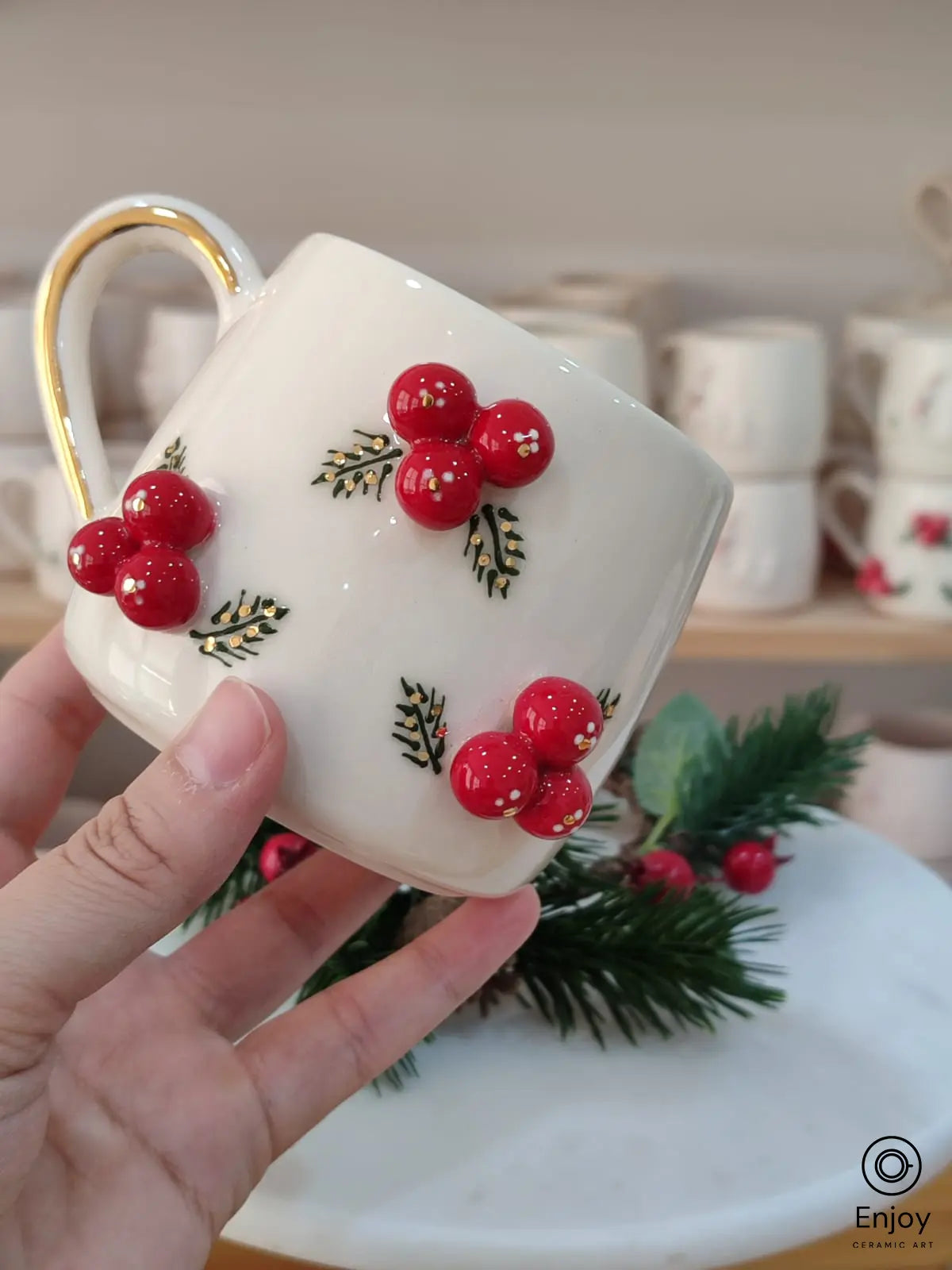 A festive handmade ceramic mug adorned with vibrant red berries and green leaf details, featuring a gold handle, perfect for adding holiday cheer to your drinkware collection.