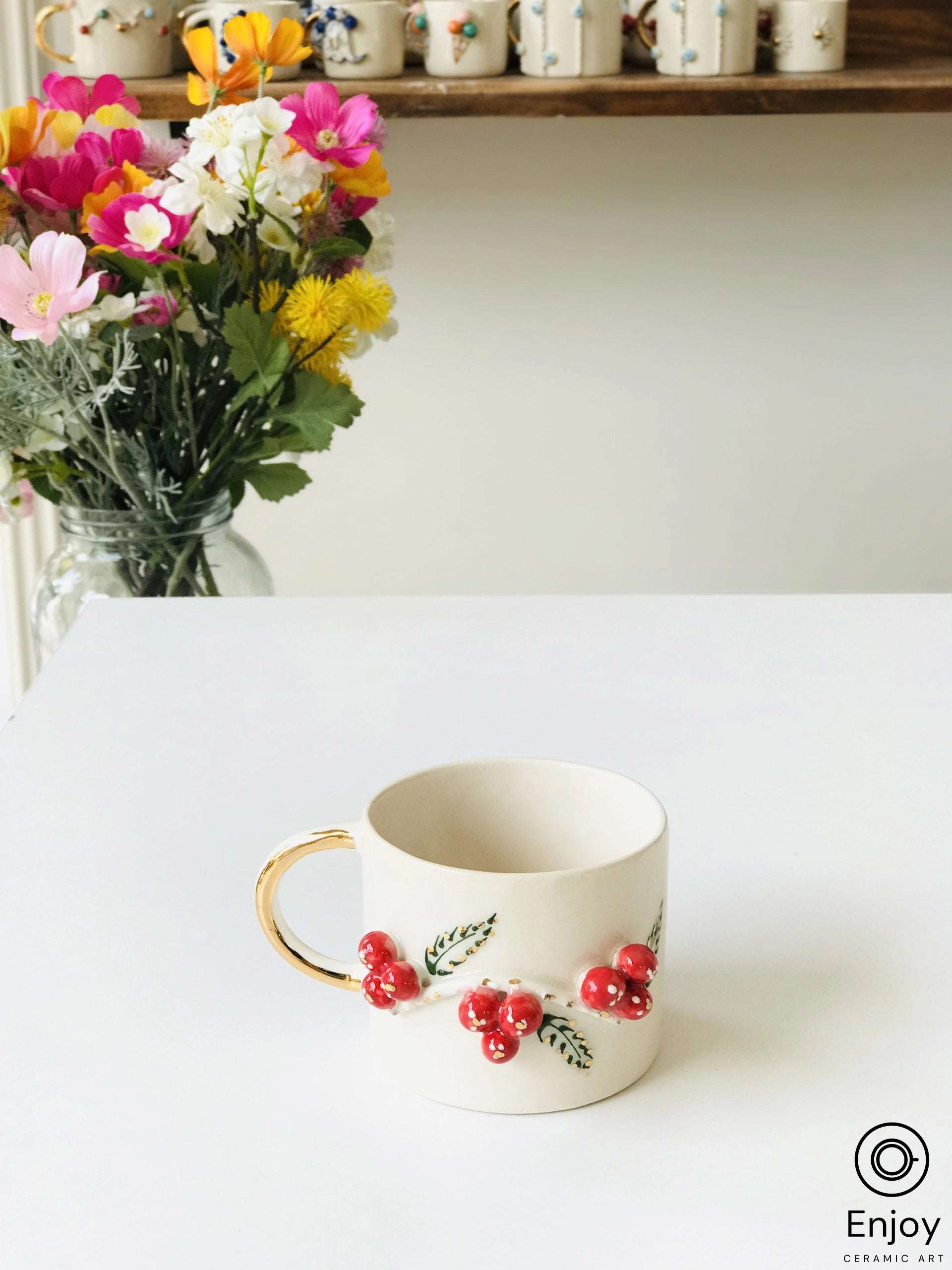 A cream-colored mug with red berry and green leaf design, golden handle, beside a vibrant bouquet on a white tabletop.