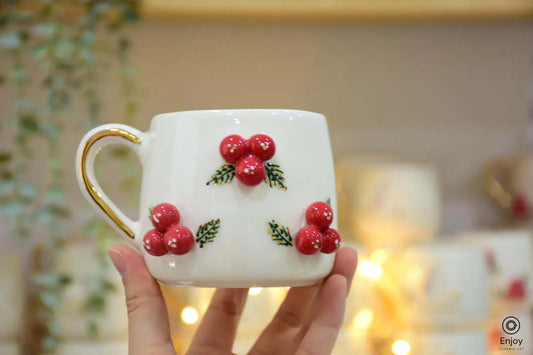 White mug with red berry decorations and golden handle, held up against a warm, festive background with soft fairy lights.