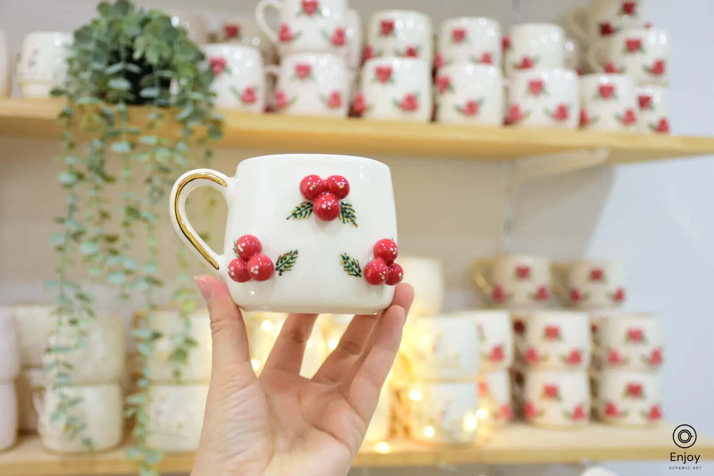 White ceramic mug with red berry decorations and a golden handle, held against a festive background with warm lighting and greenery.