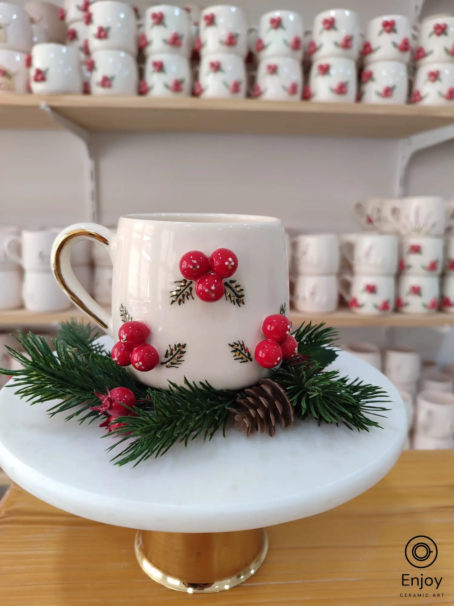 Handmade ceramic mug adorned with bright red berries and festive greenery, displayed on a marble stand with pinecone accents, perfect for holiday cheer.