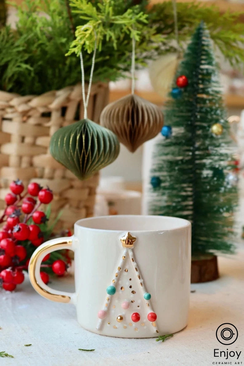 A white mug with a gold handle and a 3D Christmas tree design, surrounded by holiday decor including a basket, a small green tree, and hanging paper ornaments.