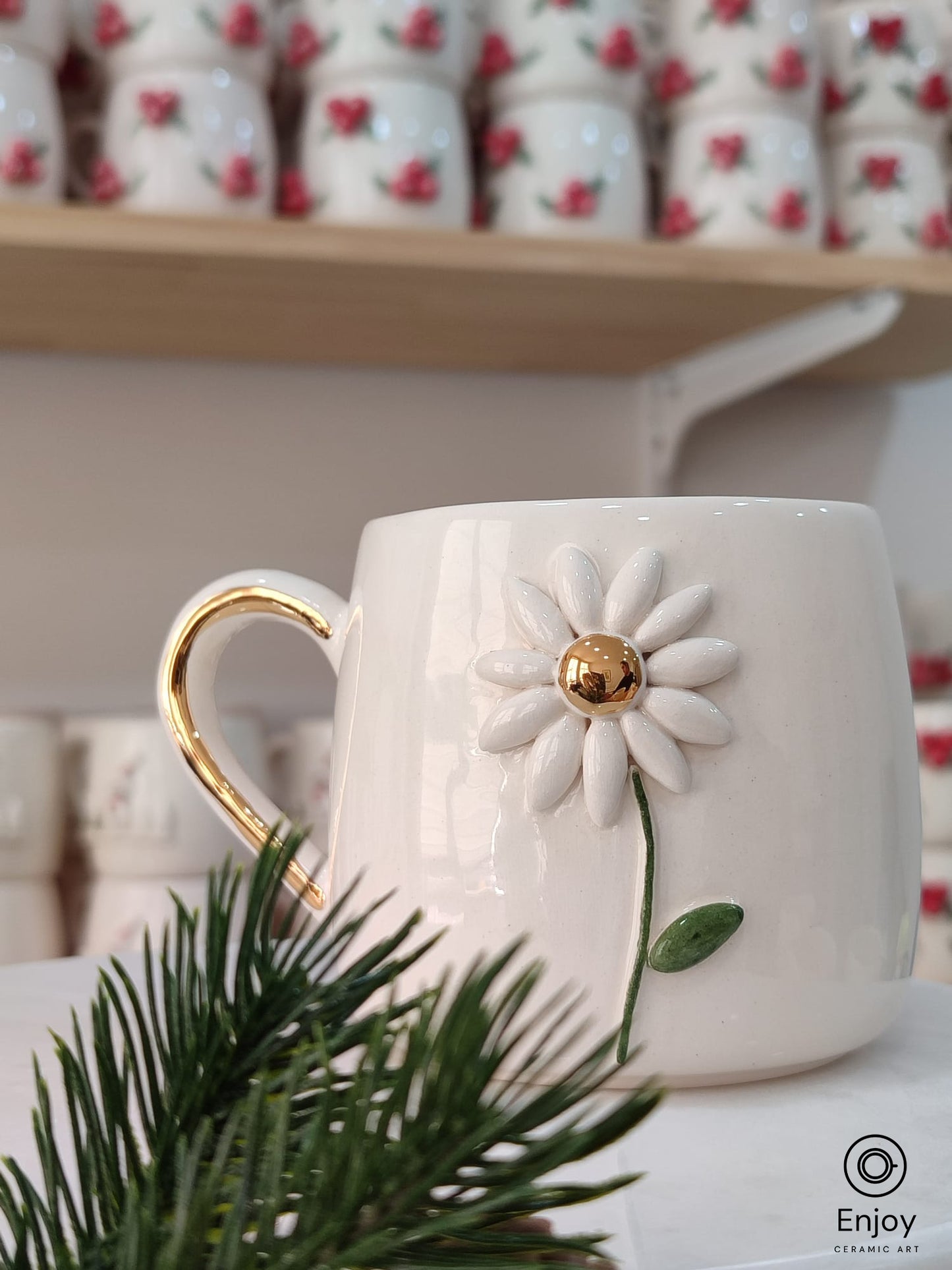 Handmade ceramic mug featuring a delicate daisy flower design with a golden center, displayed on a marble stand with festive pine decor.