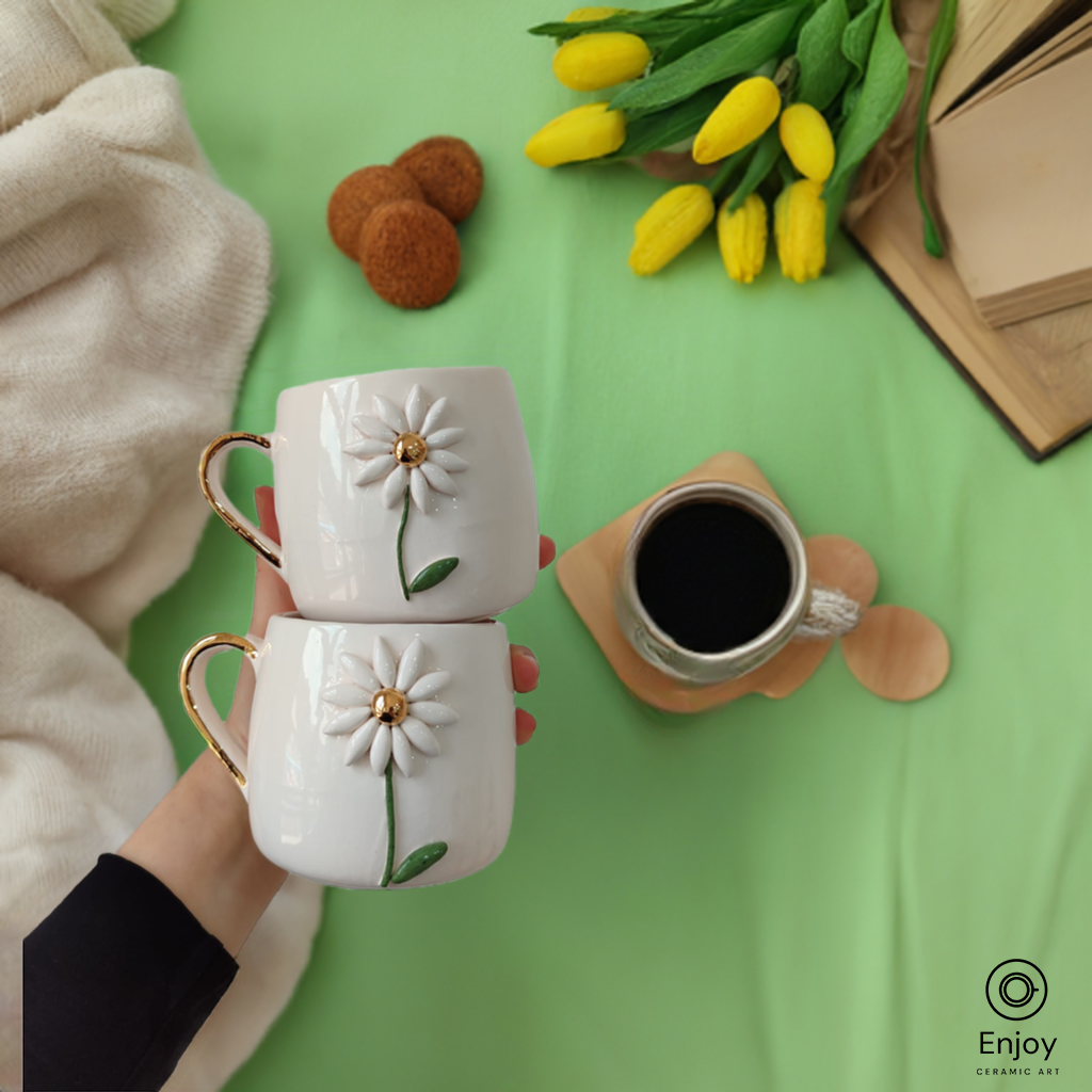 Two handmade white ceramic mugs with daisy flowers and gold handles are held together, surrounded by yellow tulips, cookies, and a cup of coffee on a green table.