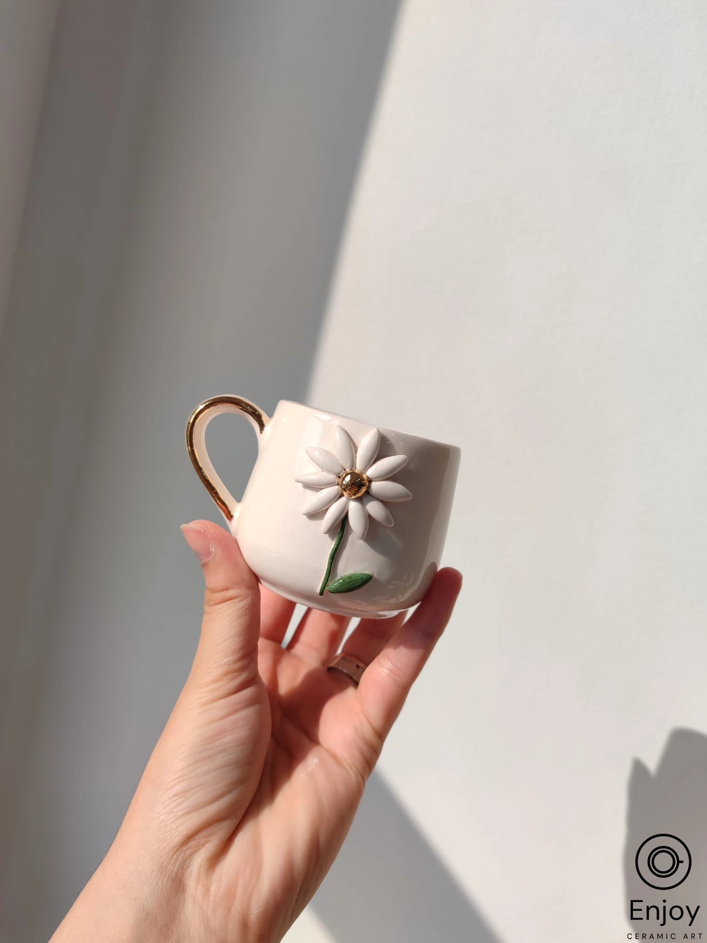 A delicate daisy-adorned mug with a gold handle, held in a hand against a shadow-cast wall.