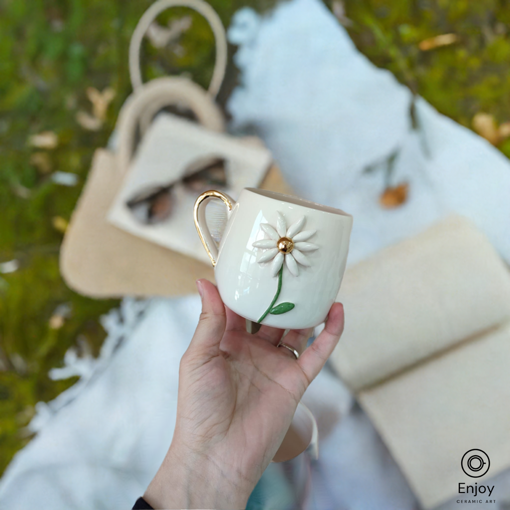 A white ceramic mug with a delicate 3D daisy design and gold handle is held outdoors, with a woven bag and blanket in the blurred background.